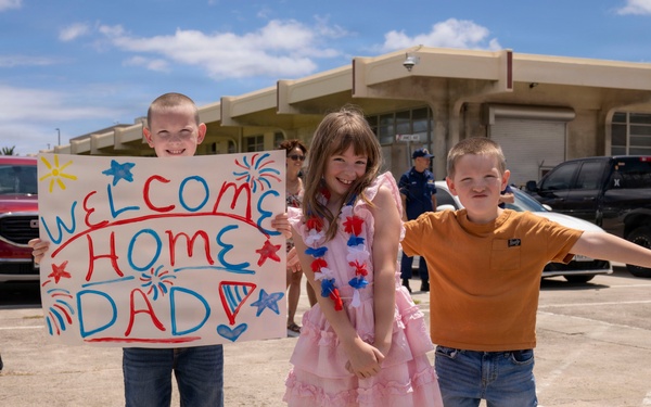 Three children stand together in a parking lot holding a handmade 'Welcome Home Dad' sign and red, white, and blue lei garlands, suggesting a military homecoming event. The children are smiling and appear excited, dressed in casual and festive attire. A uniformed Coast Guard personnel is visible in the background near a building.