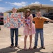 Three children stand together in a parking lot holding a handmade 'Welcome Home Dad' sign and red, white, and blue lei garlands, suggesting a military homecoming event. The children are smiling and appear excited, dressed in casual and festive attire. A uniformed Coast Guard personnel is visible in the background near a building.