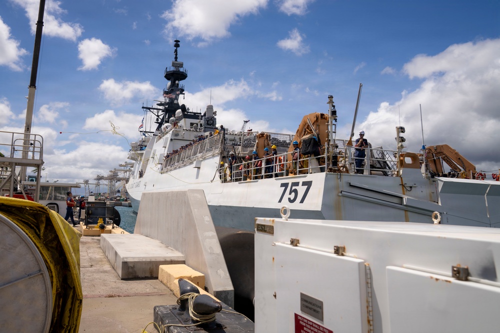 A large white Coast Guard cutter bearing hull number 757 is docked at a pier, with crew members visible on deck wearing colored hard hats. The scene appears to be a port arrival or departure operation, with personnel actively working on the vessel's deck. The setting appears to be a tropical or subtropical port based on the vegetation and weather conditions visible.