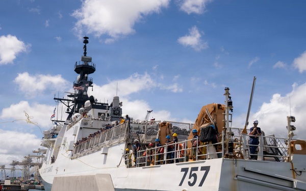 A large white Coast Guard cutter bearing hull number 757 is docked at a pier, with crew members visible on deck wearing colored hard hats. The scene appears to be a port arrival or departure operation, with personnel actively working on the vessel's deck. The setting appears to be a tropical or subtropical port based on the vegetation and weather conditions visible.