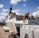 A large white Coast Guard cutter bearing hull number 757 is docked at a pier, with crew members visible on deck wearing colored hard hats. The scene appears to be a port arrival or departure operation, with personnel actively working on the vessel's deck. The setting appears to be a tropical or subtropical port based on the vegetation and weather conditions visible.