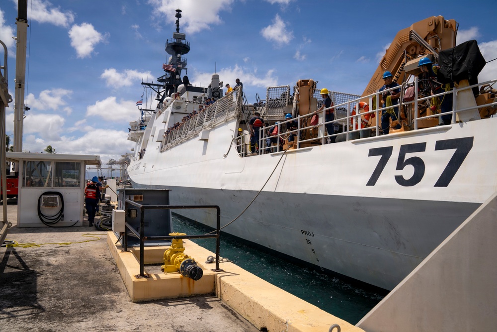 A large white military or coast guard vessel bearing hull number 757 is docked at a pier in what appears to be a tropical port. Multiple crew members wearing hard hats and safety gear are visible on deck and along the dock, suggesting active port operations or maintenance activities. Heavy equipment, including what appears to be a crane or davit system painted tan/orange, is visible on the vessel's upper deck.
