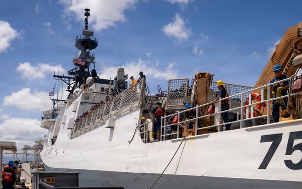 A large white military or coast guard vessel bearing hull number 757 is docked at a pier in what appears to be a tropical port. Multiple crew members wearing hard hats and safety gear are visible on deck and along the dock, suggesting active port operations or maintenance activities. Heavy equipment, including what appears to be a crane or davit system painted tan/orange, is visible on the vessel's upper deck.