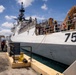 A large white military or coast guard vessel bearing hull number 757 is docked at a pier in what appears to be a tropical port. Multiple crew members wearing hard hats and safety gear are visible on deck and along the dock, suggesting active port operations or maintenance activities. Heavy equipment, including what appears to be a crane or davit system painted tan/orange, is visible on the vessel's upper deck.