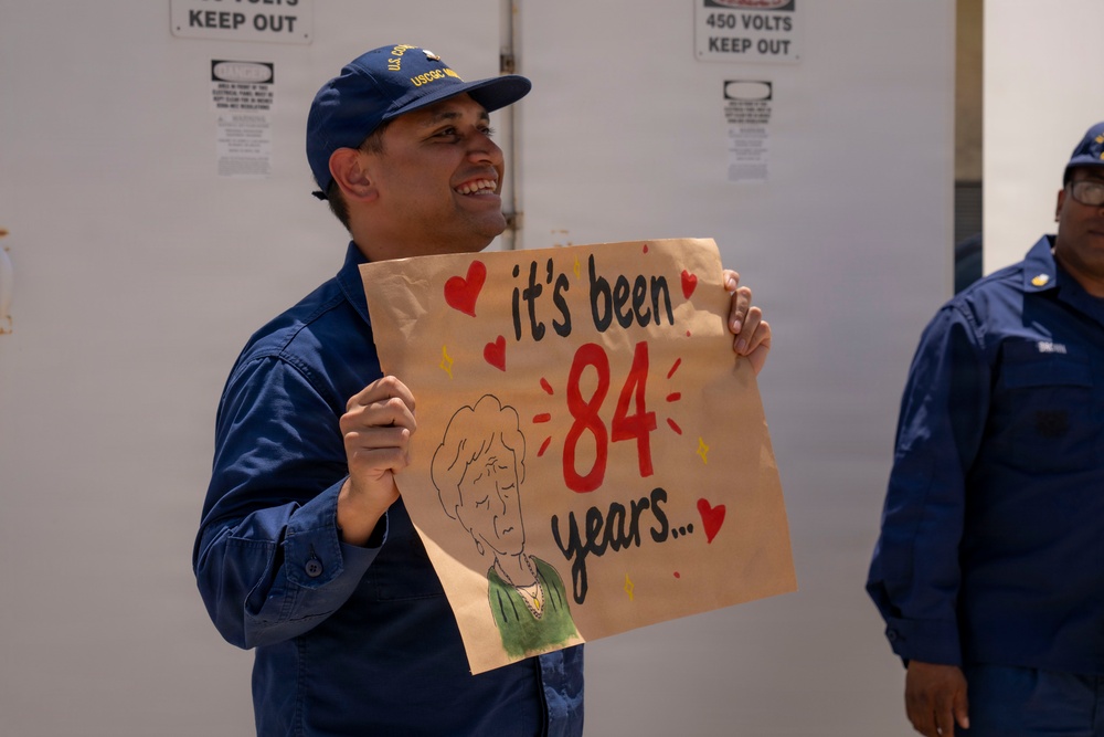 A U.S. Coast Guard member in blue uniform and USCG cap is smiling and holding a handmade sign on brown paper that reads 'it's been 84 years...' with red hearts and a cartoon drawing of an elderly woman, referencing a popular Titanic meme. A second uniformed individual is partially visible on the right side of the frame. The setting appears to be a dock or vessel area with warning signs visible in the background.