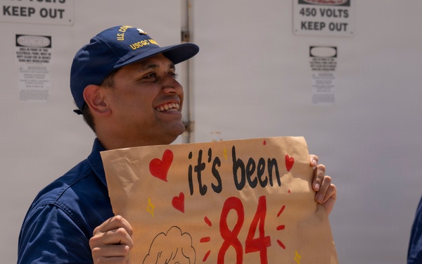 A U.S. Coast Guard member in blue uniform and USCG cap is smiling and holding a handmade sign on brown paper that reads 'it's been 84 years...' with red hearts and a cartoon drawing of an elderly woman, referencing a popular Titanic meme. A second uniformed individual is partially visible on the right side of the frame. The setting appears to be a dock or vessel area with warning signs visible in the background.