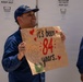 A U.S. Coast Guard member in blue uniform and USCG cap is smiling and holding a handmade sign on brown paper that reads 'it's been 84 years...' with red hearts and a cartoon drawing of an elderly woman, referencing a popular Titanic meme. A second uniformed individual is partially visible on the right side of the frame. The setting appears to be a dock or vessel area with warning signs visible in the background.