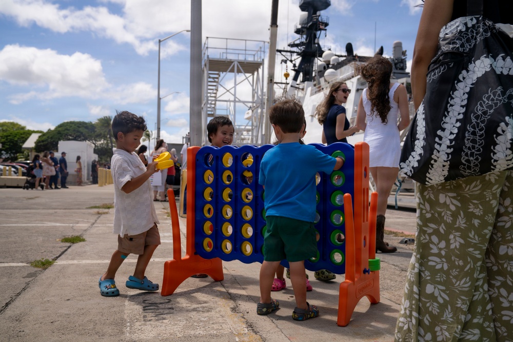 Three young children are playing with an oversized Connect Four game on a pier or dock area near a large white vessel, likely a Coast Guard or naval ship. The scene appears to be a family day or public event at a maritime facility, with adults visible in the background. The setting appears to be in a tropical or subtropical location based on the vegetation and attire.