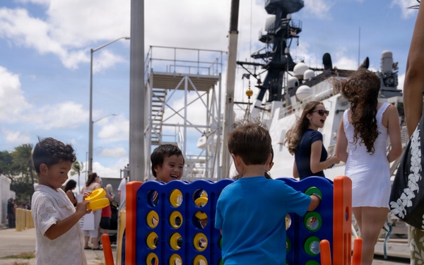 Three young children are playing with an oversized Connect Four game on a pier or dock area near a large white vessel, likely a Coast Guard or naval ship. The scene appears to be a family day or public event at a maritime facility, with adults visible in the background. The setting appears to be in a tropical or subtropical location based on the vegetation and attire.