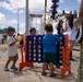 Three young children are playing with an oversized Connect Four game on a pier or dock area near a large white vessel, likely a Coast Guard or naval ship. The scene appears to be a family day or public event at a maritime facility, with adults visible in the background. The setting appears to be in a tropical or subtropical location based on the vegetation and attire.
