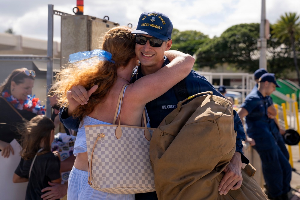 A U.S. Coast Guard member assigned to the USCGC Midgett is embracing a woman with red hair in what appears to be a homecoming or departure event at a port facility. The servicemember is smiling broadly while holding a duffel bag, suggesting a reunion after deployment. Other Coast Guard personnel and civilians are visible in the background.