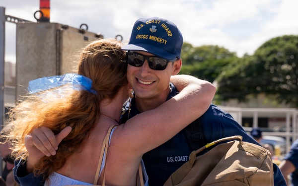 A U.S. Coast Guard member assigned to the USCGC Midgett is embracing a woman with red hair in what appears to be a homecoming or departure event at a port facility. The servicemember is smiling broadly while holding a duffel bag, suggesting a reunion after deployment. Other Coast Guard personnel and civilians are visible in the background.