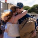 A U.S. Coast Guard member assigned to the USCGC Midgett is embracing a woman with red hair in what appears to be a homecoming or departure event at a port facility. The servicemember is smiling broadly while holding a duffel bag, suggesting a reunion after deployment. Other Coast Guard personnel and civilians are visible in the background.
