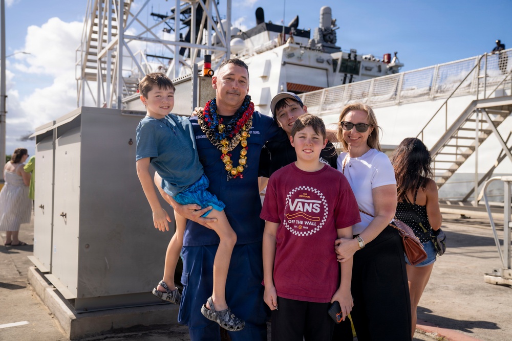 A U.S. Coast Guard service member wearing a lei is reuniting with his family at a dock, likely following a deployment. The group of five — including what appear to be three children and a woman — pose together in front of a large white Coast Guard vessel. The setting suggests a homecoming ceremony at a port, possibly in Hawaii given the tropical environment and lei.