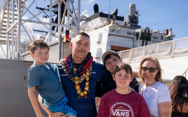 A U.S. Coast Guard service member wearing a lei is reuniting with his family at a dock, likely following a deployment. The group of five — including what appear to be three children and a woman — pose together in front of a large white Coast Guard vessel. The setting suggests a homecoming ceremony at a port, possibly in Hawaii given the tropical environment and lei.