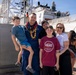 A U.S. Coast Guard service member wearing a lei is reuniting with his family at a dock, likely following a deployment. The group of five — including what appear to be three children and a woman — pose together in front of a large white Coast Guard vessel. The setting suggests a homecoming ceremony at a port, possibly in Hawaii given the tropical environment and lei.