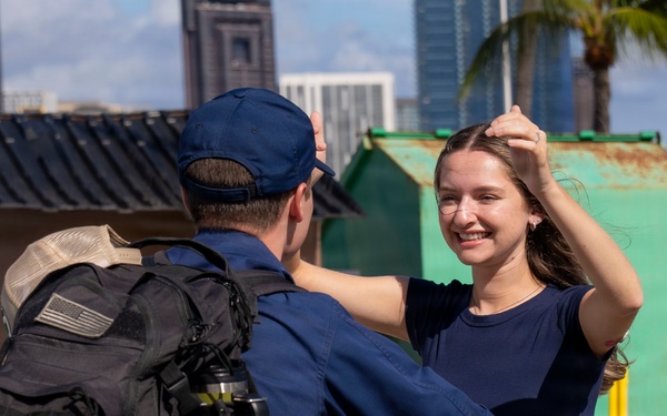 A man in a dark blue uniform and black tactical backpack is greeting or embracing a smiling young woman in a navy blue shirt and jeans. The setting appears to be a port or waterfront area with urban buildings and a palm tree visible in the background. The interaction suggests a reunion, possibly a military or Coast Guard homecoming.