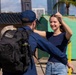A man in a dark blue uniform and black tactical backpack is greeting or embracing a smiling young woman in a navy blue shirt and jeans. The setting appears to be a port or waterfront area with urban buildings and a palm tree visible in the background. The interaction suggests a reunion, possibly a military or Coast Guard homecoming.