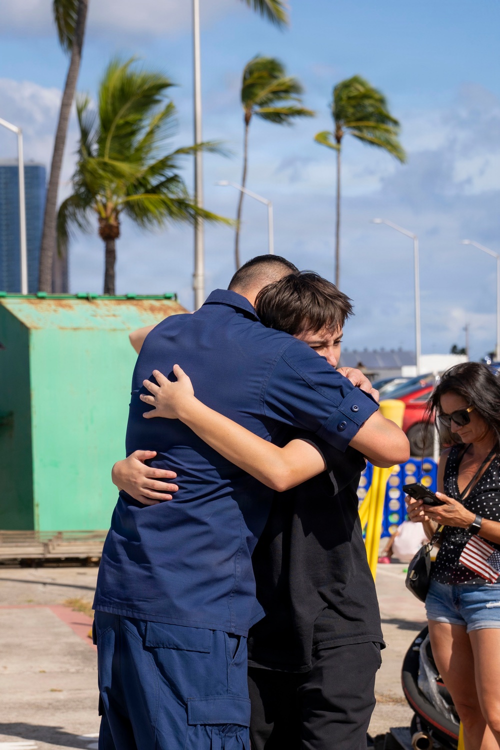 Two individuals are sharing an emotional embrace in an outdoor coastal setting with palm trees swaying in the wind. One person is wearing a navy blue uniform consistent with military or Coast Guard attire, suggesting this may be a homecoming or farewell event. A woman holding a small American flag stands nearby, further supporting the context of a military-related gathering.