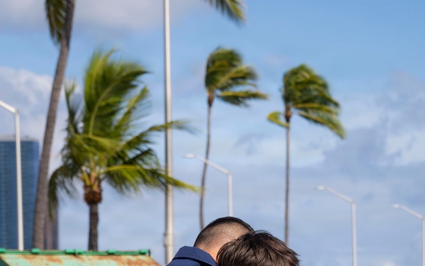 Two individuals are sharing an emotional embrace in an outdoor coastal setting with palm trees swaying in the wind. One person is wearing a navy blue uniform consistent with military or Coast Guard attire, suggesting this may be a homecoming or farewell event. A woman holding a small American flag stands nearby, further supporting the context of a military-related gathering.