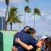 Two individuals are sharing an emotional embrace in an outdoor coastal setting with palm trees swaying in the wind. One person is wearing a navy blue uniform consistent with military or Coast Guard attire, suggesting this may be a homecoming or farewell event. A woman holding a small American flag stands nearby, further supporting the context of a military-related gathering.