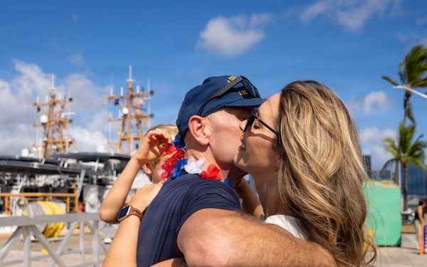 A military homecoming scene at a naval pier, where a man in a Navy cap is kissing a woman while holding a young child. A second child wearing a red, white, and blue lei stands in the background smiling. The setting appears to be a tropical port, likely in Hawaii based on the palm trees and urban skyline.