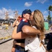 A military homecoming scene at a naval pier, where a man in a Navy cap is kissing a woman while holding a young child. A second child wearing a red, white, and blue lei stands in the background smiling. The setting appears to be a tropical port, likely in Hawaii based on the palm trees and urban skyline.