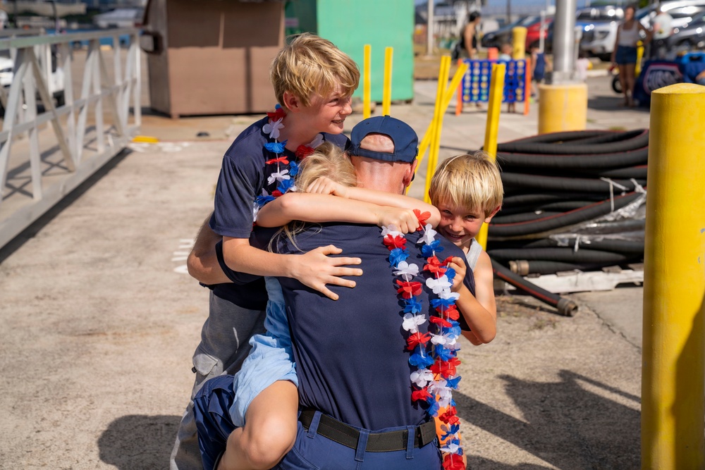 An emotional reunion scene showing an adult in a navy blue uniform and cap being embraced by three blonde children wearing red, white, and blue lei garlands. The setting appears to be a military or naval pier/dock facility, suggesting this may be a homecoming event. The children are visibly joyful, with one older boy hugging from behind, a younger child in front, and a third child being held.