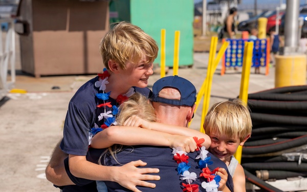 An emotional reunion scene showing an adult in a navy blue uniform and cap being embraced by three blonde children wearing red, white, and blue lei garlands. The setting appears to be a military or naval pier/dock facility, suggesting this may be a homecoming event. The children are visibly joyful, with one older boy hugging from behind, a younger child in front, and a third child being held.