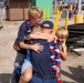 An emotional reunion scene showing an adult in a navy blue uniform and cap being embraced by three blonde children wearing red, white, and blue lei garlands. The setting appears to be a military or naval pier/dock facility, suggesting this may be a homecoming event. The children are visibly joyful, with one older boy hugging from behind, a younger child in front, and a third child being held.