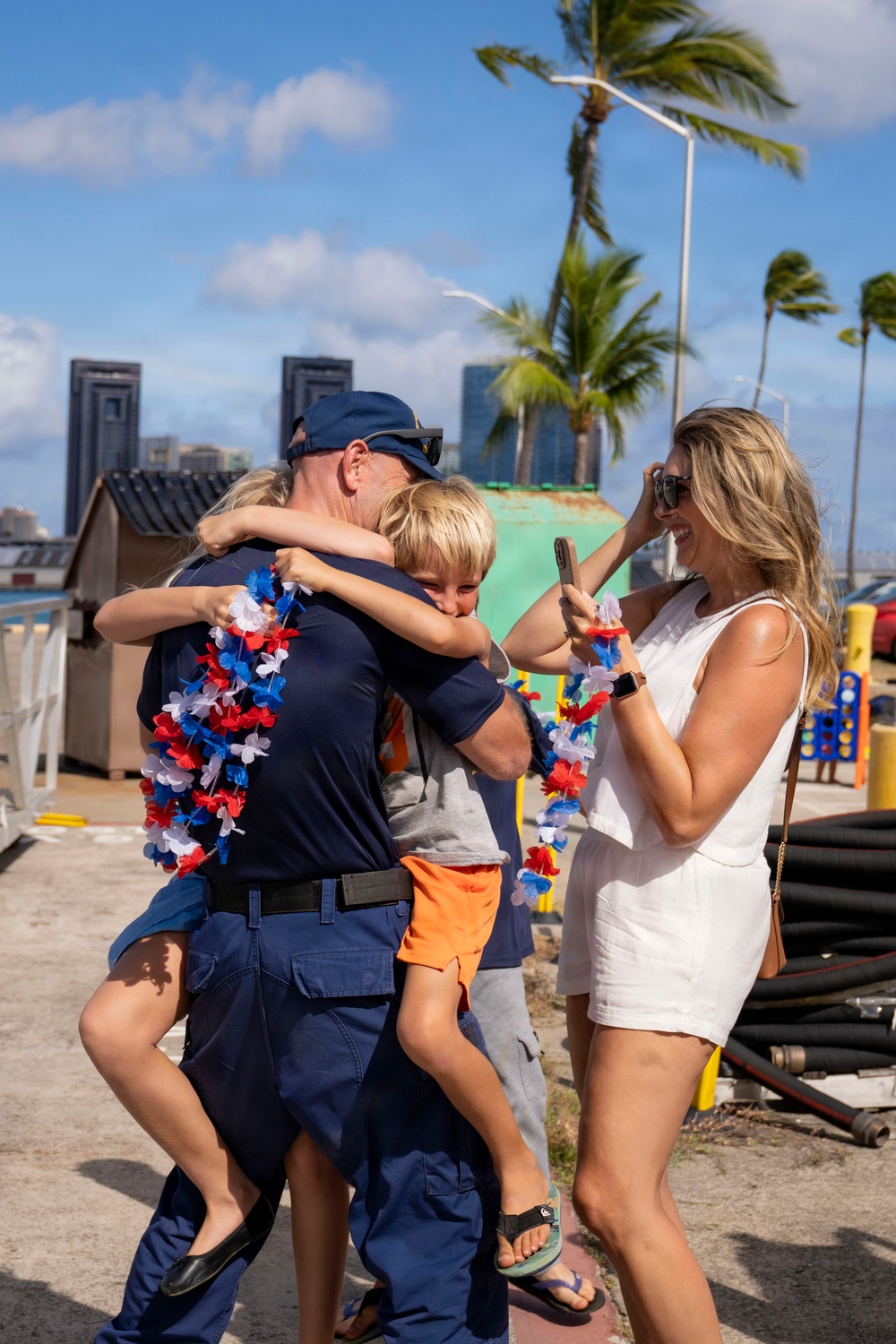 A joyful reunion scene at what appears to be a harbor or port facility in a tropical location, likely Hawaii, based on the palm trees and urban skyline in the background. A man in navy blue uniform with red, white, and blue leis is embracing two children while a blonde woman in a white dress photographs the moment on her phone. The scene strongly suggests a military or Coast Guard homecoming.