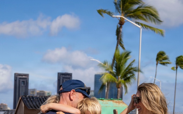A joyful reunion scene at what appears to be a harbor or port facility in a tropical location, likely Hawaii, based on the palm trees and urban skyline in the background. A man in navy blue uniform with red, white, and blue leis is embracing two children while a blonde woman in a white dress photographs the moment on her phone. The scene strongly suggests a military or Coast Guard homecoming.