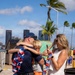 A joyful reunion scene at what appears to be a harbor or port facility in a tropical location, likely Hawaii, based on the palm trees and urban skyline in the background. A man in navy blue uniform with red, white, and blue leis is embracing two children while a blonde woman in a white dress photographs the moment on her phone. The scene strongly suggests a military or Coast Guard homecoming.