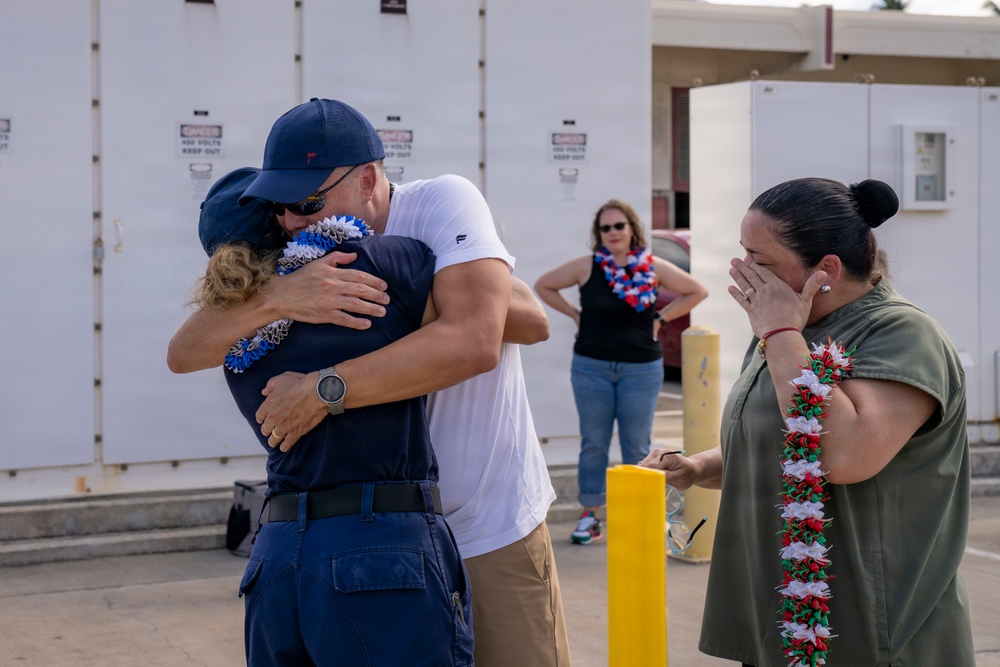 An emotional reunion scene showing people embracing and crying, likely at a military or official homecoming event. Multiple individuals are wearing decorative leis, suggesting a Hawaiian or Pacific location. A woman on the right is visibly emotional, covering her face while crying.