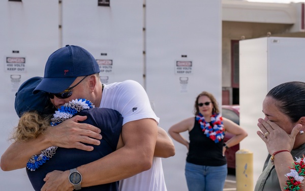 An emotional reunion scene showing people embracing and crying, likely at a military or official homecoming event. Multiple individuals are wearing decorative leis, suggesting a Hawaiian or Pacific location. A woman on the right is visibly emotional, covering her face while crying.