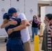 An emotional reunion scene showing people embracing and crying, likely at a military or official homecoming event. Multiple individuals are wearing decorative leis, suggesting a Hawaiian or Pacific location. A woman on the right is visibly emotional, covering her face while crying.