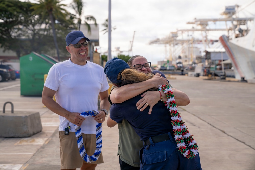 A warm reunion scene at a port or harbor, where two people are embracing in an emotional hug. One person is wearing a red, white, and green lei while another man stands nearby smiling and holding a blue and white lei. The setting appears to be a working waterfront with cranes and vessels visible in the background.