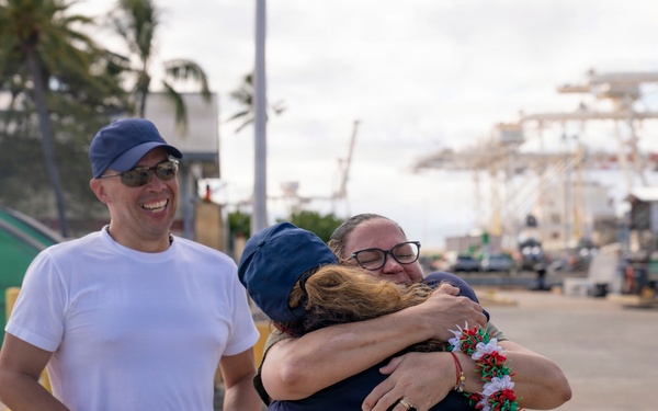 A warm reunion scene at a port or harbor, where two people are embracing in an emotional hug. One person is wearing a red, white, and green lei while another man stands nearby smiling and holding a blue and white lei. The setting appears to be a working waterfront with cranes and vessels visible in the background.