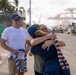A warm reunion scene at a port or harbor, where two people are embracing in an emotional hug. One person is wearing a red, white, and green lei while another man stands nearby smiling and holding a blue and white lei. The setting appears to be a working waterfront with cranes and vessels visible in the background.