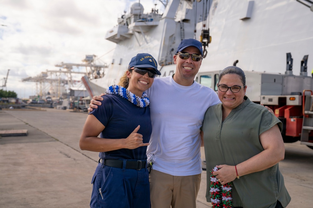 Three smiling individuals pose together on a dock in front of a large white U.S. Coast Guard vessel. The person on the left is wearing a U.S. Coast Guard uniform and cap with a blue and white lei, while the person on the right holds a red and white lei, suggesting a homecoming or departure ceremony. The setting appears to be a military or commercial port facility.