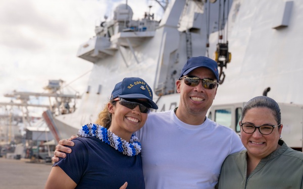 Three smiling individuals pose together on a dock in front of a large white U.S. Coast Guard vessel. The person on the left is wearing a U.S. Coast Guard uniform and cap with a blue and white lei, while the person on the right holds a red and white lei, suggesting a homecoming or departure ceremony. The setting appears to be a military or commercial port facility.
