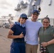 Three smiling individuals pose together on a dock in front of a large white U.S. Coast Guard vessel. The person on the left is wearing a U.S. Coast Guard uniform and cap with a blue and white lei, while the person on the right holds a red and white lei, suggesting a homecoming or departure ceremony. The setting appears to be a military or commercial port facility.