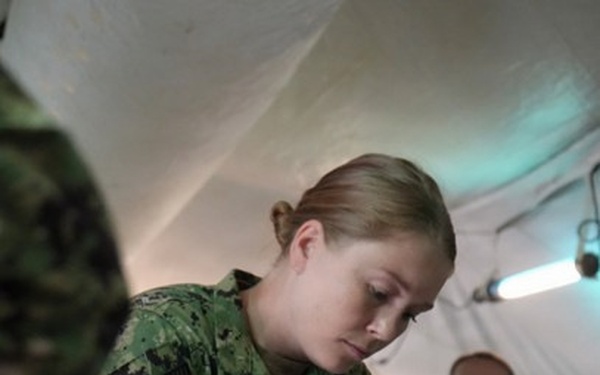 A U.S. Navy service member named 'Capps' is performing a medical procedure or documentation task while wearing blue nitrile gloves inside what appears to be a military field medical tent. A second uniformed service member wearing glasses observes in the background. The setting suggests a field medical training exercise or operational medical environment.