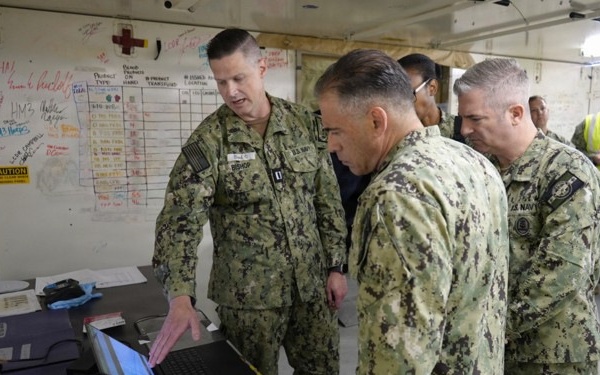 Several U.S. Navy personnel in NWU Type III camouflage uniforms are gathered around a laptop computer in what appears to be a military operations or medical briefing area. One officer, whose name tape reads 'BISHOP,' is pointing at the laptop screen while presenting information to the others. A whiteboard with tracking charts, medical symbols, and handwritten notes is visible in the background.