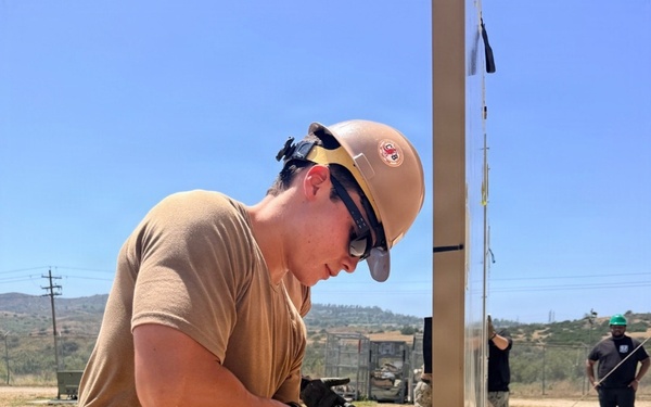 A military service member wearing Navy Working Uniform (NWU) camouflage and a tan hard hat is performing construction work, using a hammer or mallet on a metal structural frame. The setting appears to be an outdoor construction site in an arid, hilly environment, likely a military installation. Additional personnel are visible in the background, including one individual wearing a green hard hat.