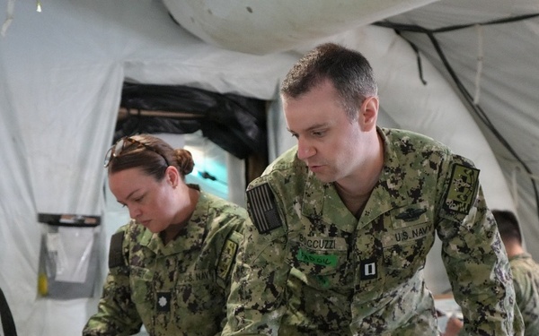 Two U.S. Navy personnel in AOR2 digital camouflage uniforms are working at a table inside a military field tent or temporary structure. The foreground individual, whose name tape reads 'ACCUZZI' and bears a Captain's rank insignia, is leaning over a laptop computer. A third service member is visible seated in the background.