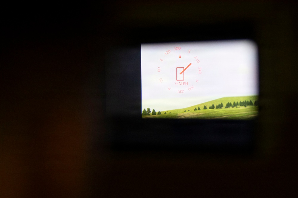 The image shows a heads-up display (HUD) or augmented reality overlay visible through a small rectangular viewport or eyepiece. The HUD displays navigational data including a compass rose with degree markings and a speed readout of 0 MPH, overlaid on a simulated or real outdoor scene featuring green rolling hills and conifer trees. The surrounding area is dark, suggesting the viewer is looking through a device such as a scope, viewfinder, or simulator display.