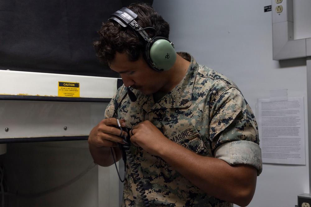 A U.S. Marine in MARPAT digital camouflage uniform is wearing green hearing protection headphones and handling a coiled cable or communication device. He appears to be in an indoor facility, possibly a shooting range or technical workspace. The 'MARINES' name tape is clearly visible on his uniform.