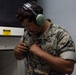 A U.S. Marine in MARPAT digital camouflage uniform is wearing green hearing protection headphones and handling a coiled cable or communication device. He appears to be in an indoor facility, possibly a shooting range or technical workspace. The 'MARINES' name tape is clearly visible on his uniform.