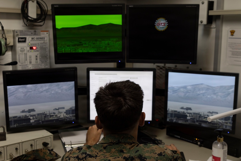 A military personnel member in camouflage uniform is seated at a multi-monitor workstation, appearing to operate a surveillance or targeting simulation system. The setup includes five NEC monitors displaying various feeds including a green night-vision-style view, daylight camera views of a mountainous terrain with structures, and a text interface. A logo reading 'BTS' is visible on one of the upper monitors, suggesting a training or simulation system.